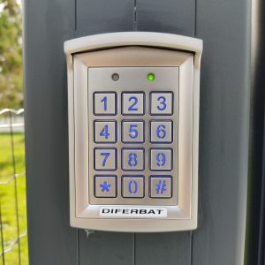 Un clavier numérique extérieur de marque Diferbat avec une lumière verte, fixé sur un portail gris, dans un cadre de jardin.