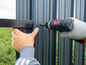 Un homme utilise une perceuse pour assembler un portail métallique gris, avec une main tenant la structure et un fond de verdure.
