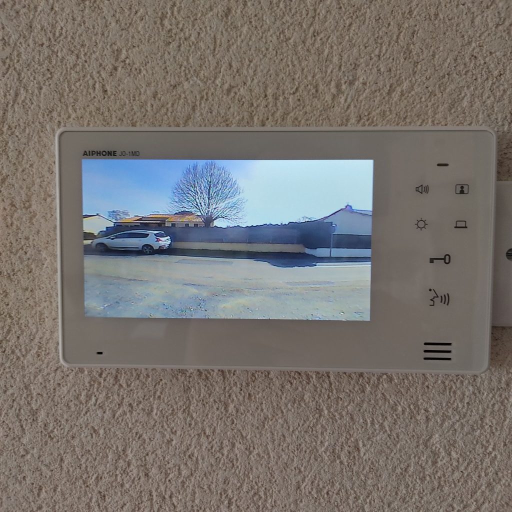 Un interphone vidéo blanc de marque Aiphone affichant une image d'un extérieur avec une voiture et une maison, monté sur un mur texturé.