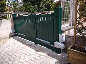 Un portail vert à lames verticales avec un design courbé, encadré par un mur en pierre, sur une cour pavée avec des moteurs automatiques visibles.