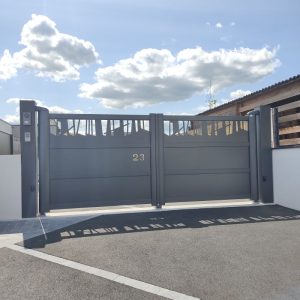Un portail double gris métallique avec des motifs découpés en forme d'arbres, numéroté "23", situé devant une maison avec un interphone sur le côté, sous un ciel nuageux.