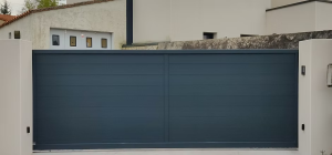 Un large portail gris foncé à panneaux lisses, encadré par des murs blancs, devant une maison avec un toit en tuiles rouges, sur une entrée pavée.