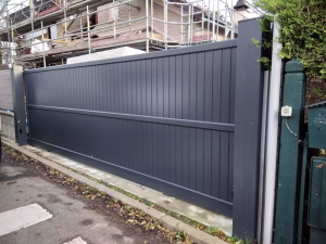Un portail gris foncé à lames verticales, installé devant une maison en construction avec des échafaudages, sur un trottoir bordé de végétation.
