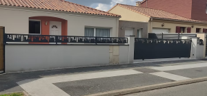 Une maison avec un toit en tuiles rouges et une façade beige, entourée d'un mur avec un portail et une clôture noirs ornés de motifs, donnant sur une rue avec un passage piéton.