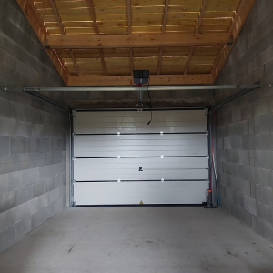 Une porte de garage blanche à panneaux horizontaux, montée dans une structure en parpaings avec un toit en bois, équipée d’un mécanisme d’ouverture automatique.