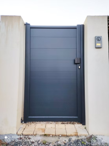 Un portillon d'entrée gris à panneaux horizontaux, avec une serrure et un interphone à droite, encastré dans un mur clair.