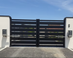 Un portail noir à lames horizontales, encadré par des murs blancs, sur une entrée pavée, avec des moteurs automatiques de chaque côté, sous un ciel bleu.